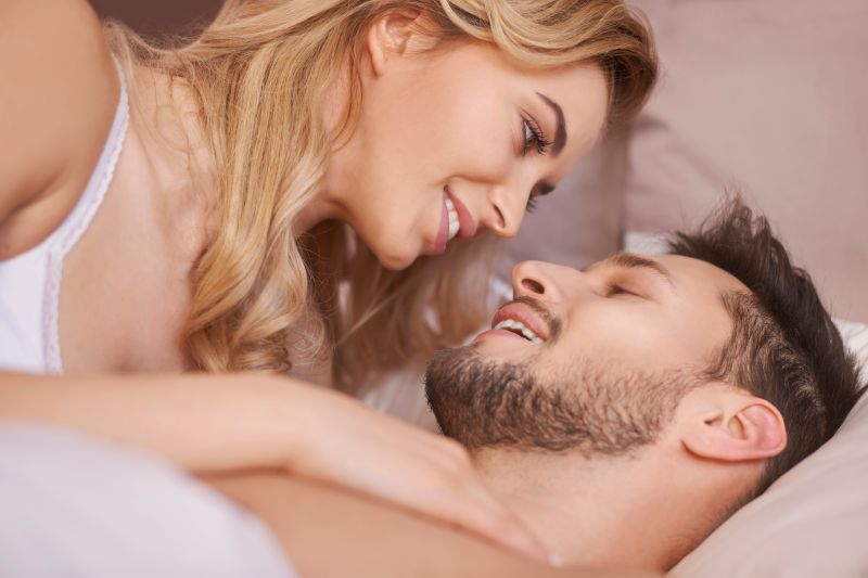 Brunette bearded man lying below blond woman with hand on man's chest. Both smiling